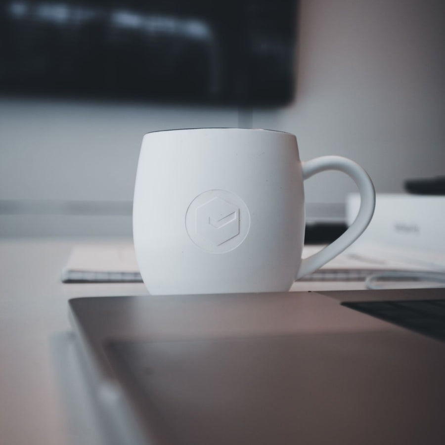 A white mug photographed on an office desk in a plain minimal setting.