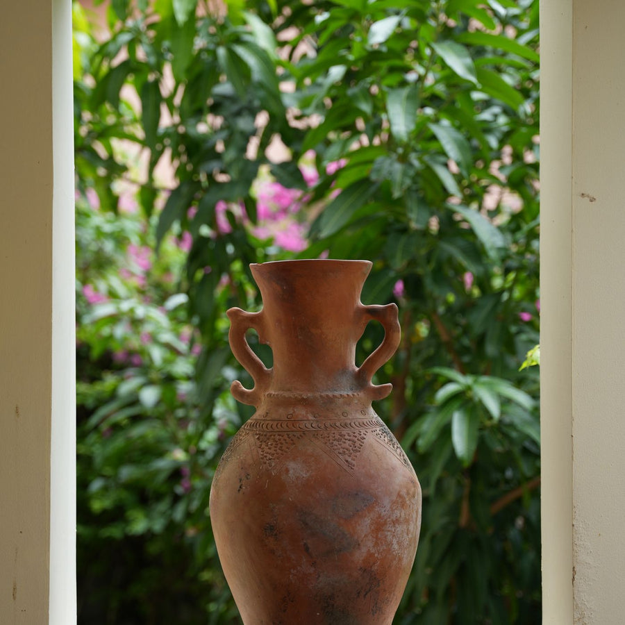 A rustic clay vase set in a window with greenery framing the opening.