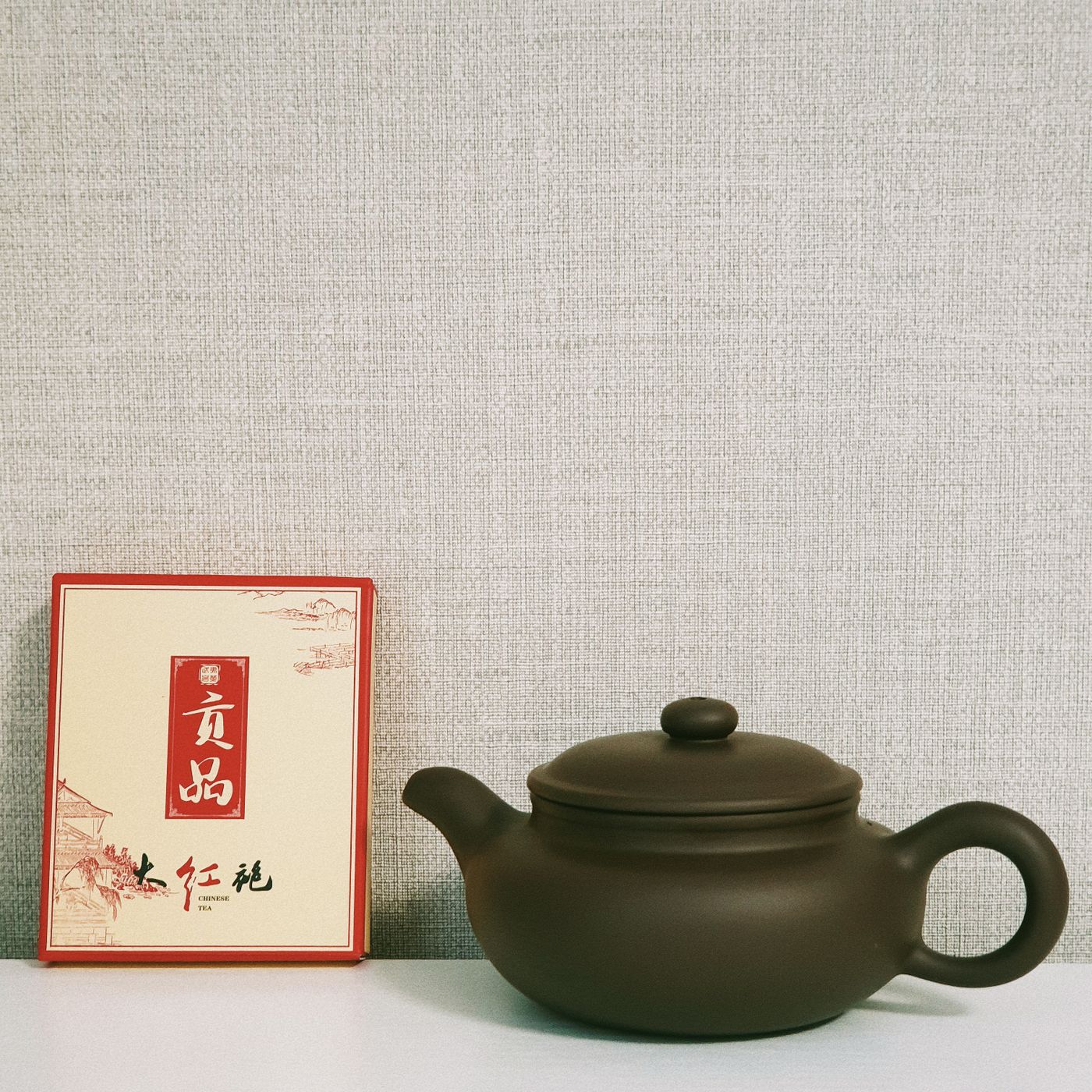 A small clay teapot photographed on a plain background beside an oriental themed box.