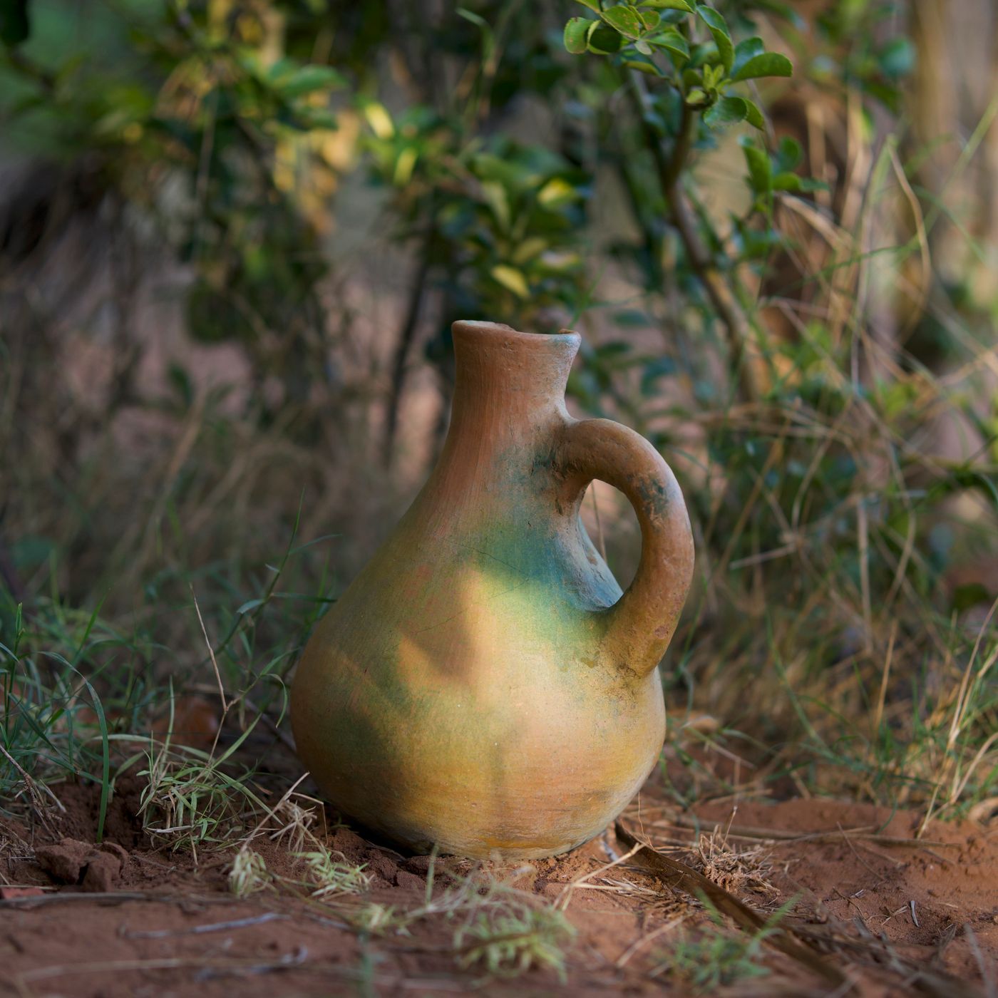 A rustic clay jug standing on bare ground with green foliage around it.