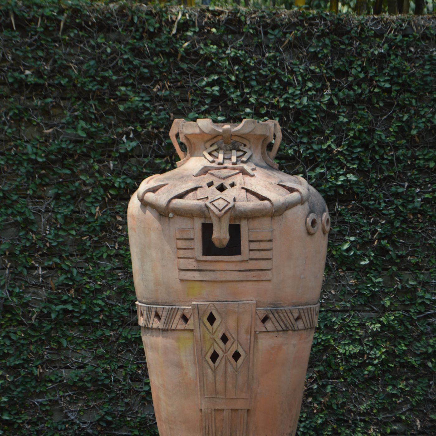 A clay vase worked with intricate designs, shown outdoors on a stone pedestal.