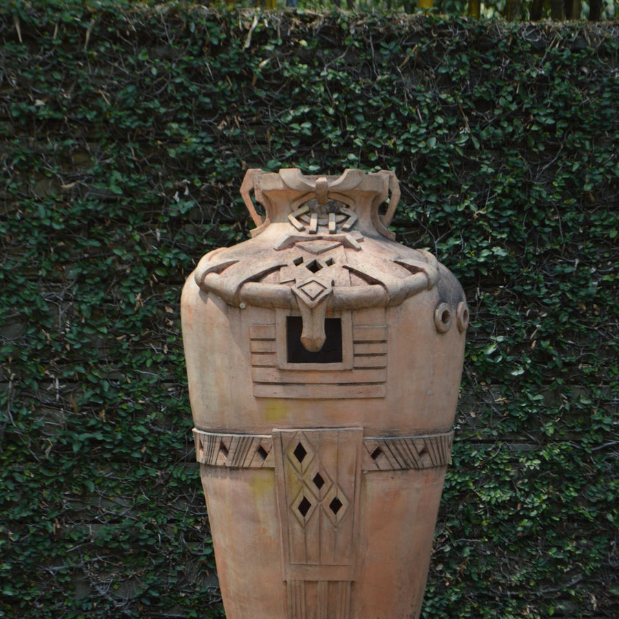 A clay vase worked with intricate designs, shown outdoors on a stone pedestal.