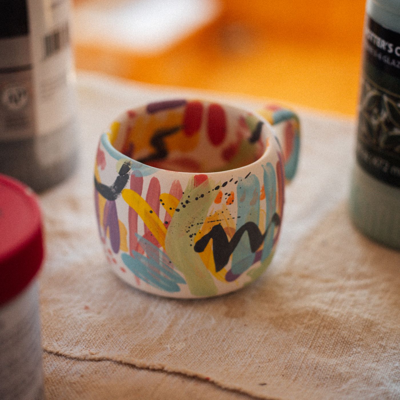 A hand painted ceramic mug in vivid colours, shown on a textured surface with paint bottles around it.