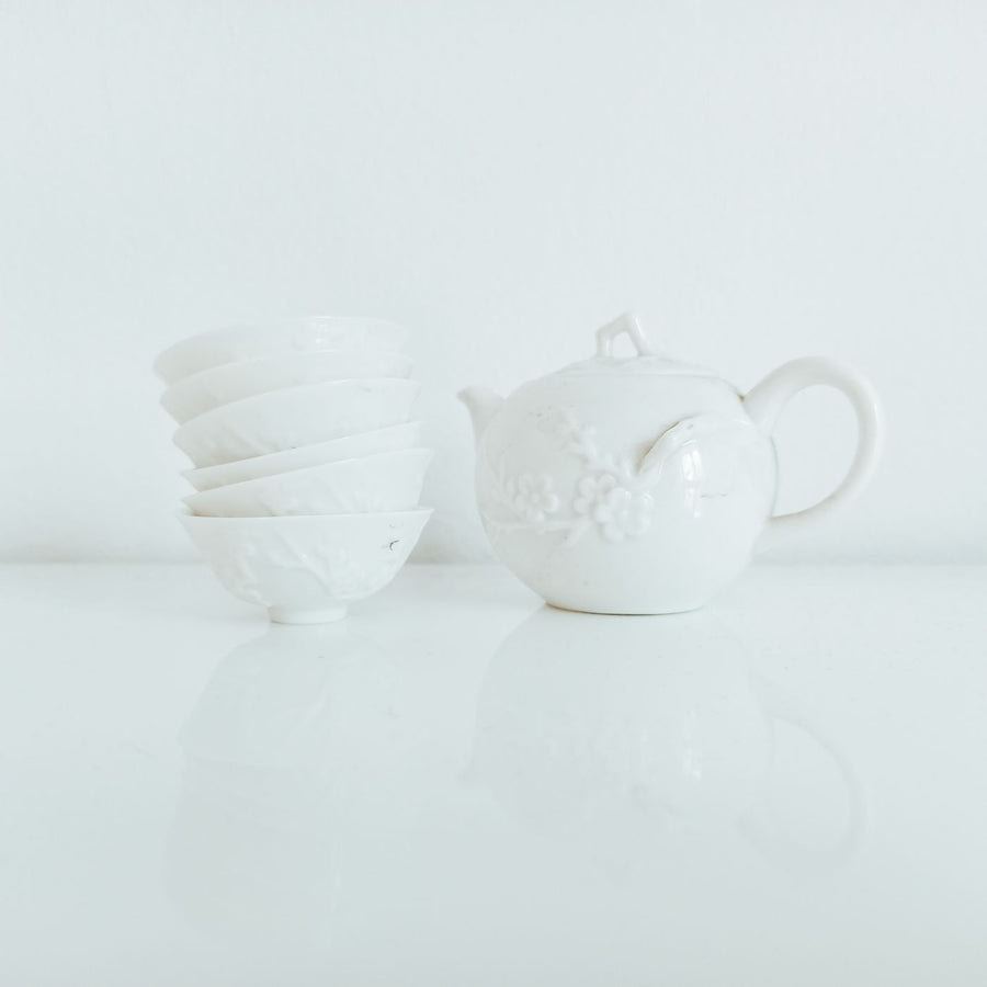 A white ceramic teapot in an old fashioned shape, set on a polished table near chawan bowls.