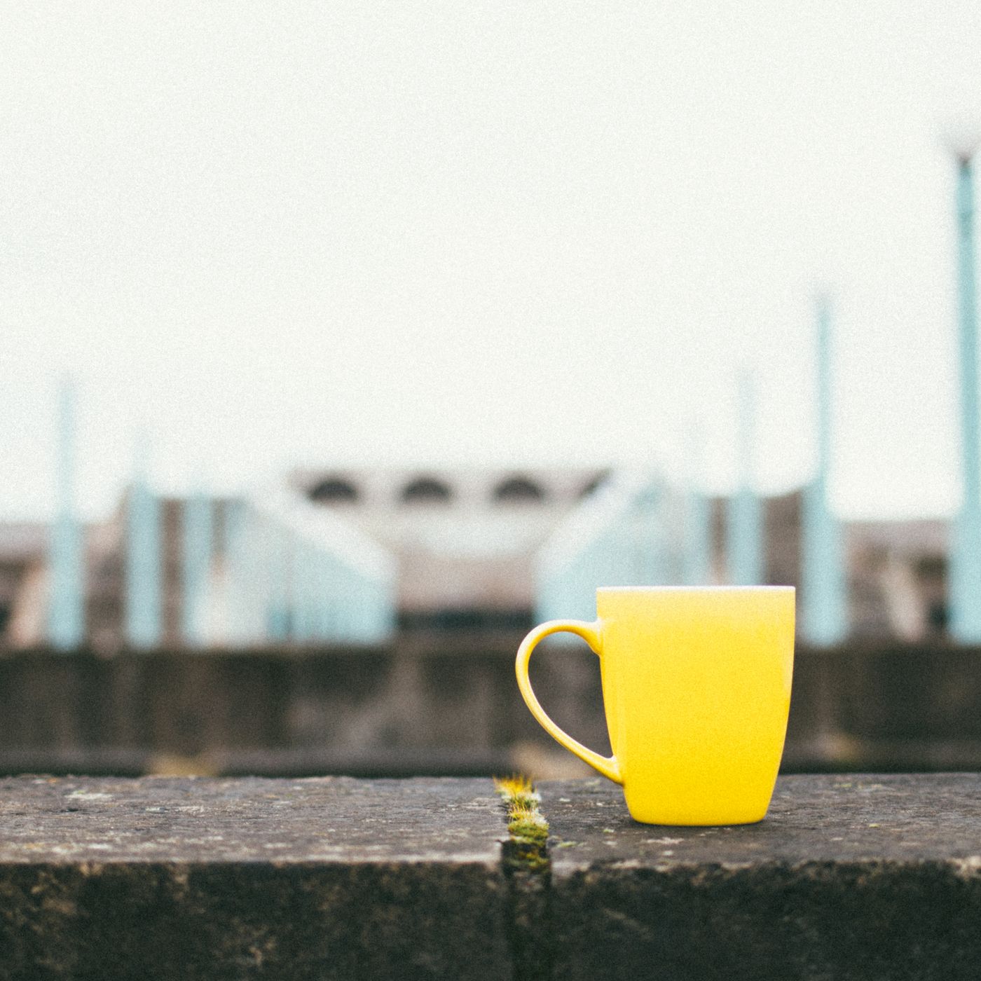 A bright yellow mug set down on concrete, with blurred architecture behind it.