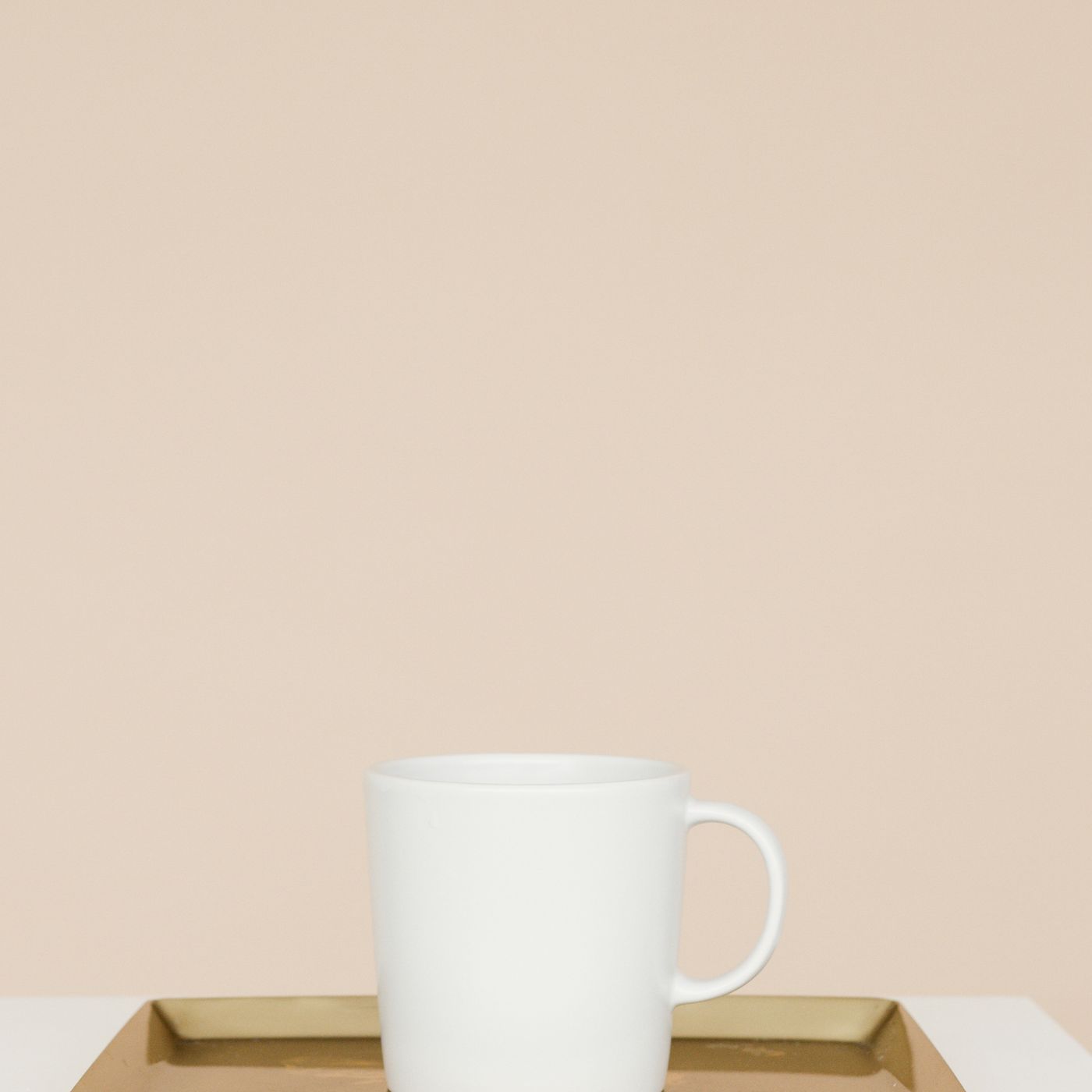 A simple white ceramic mug standing on a gold tray against a beige background.