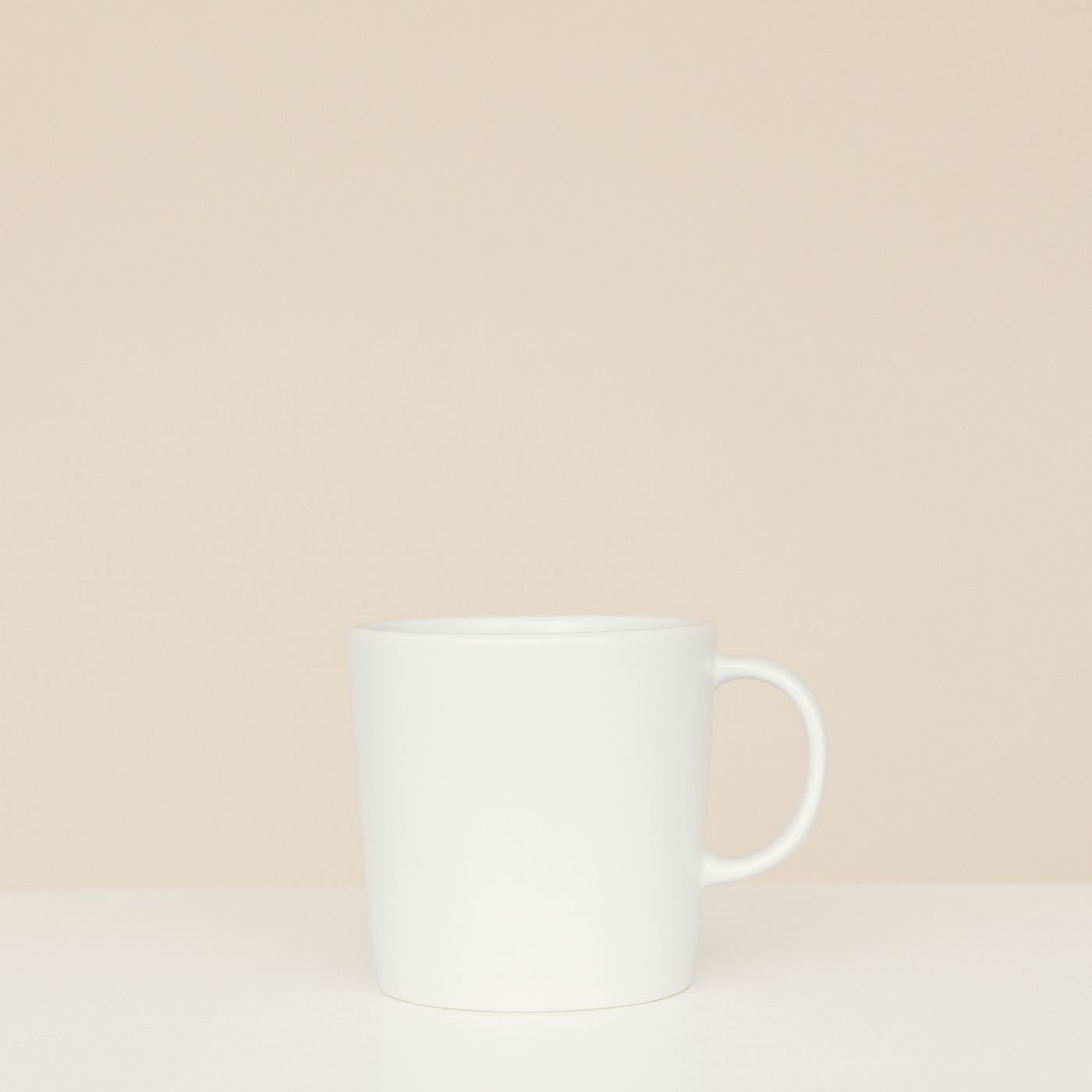 A simple white ceramic mug shown on a neutral beige background with nothing else in frame.