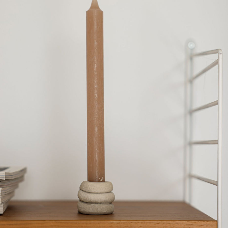 A clay candle holder on a plain wooden shelf with a tall brown candle standing in it.