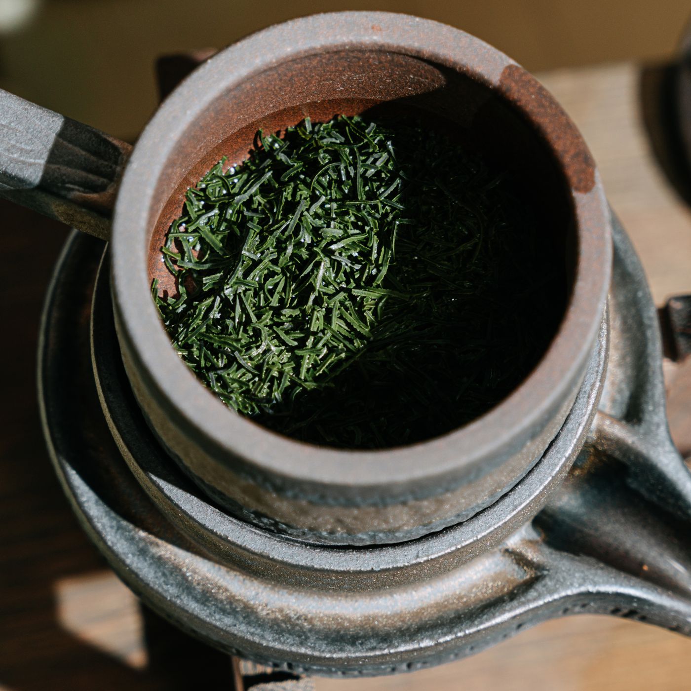 A rustic clay teapot photographed close up holding dry green tea leaves.