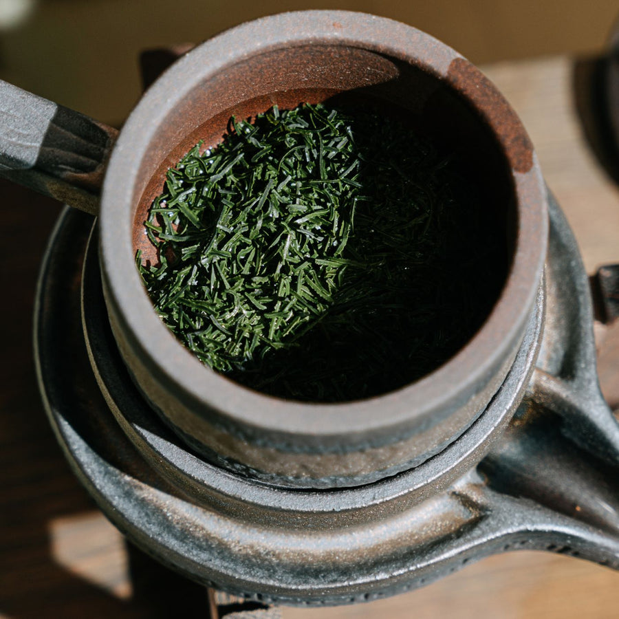 A rustic clay teapot photographed close up holding dry green tea leaves.